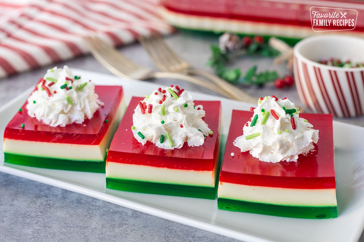 Three slices of Christmas Jell-O salad on a plate with whipped cream and sprinkles.