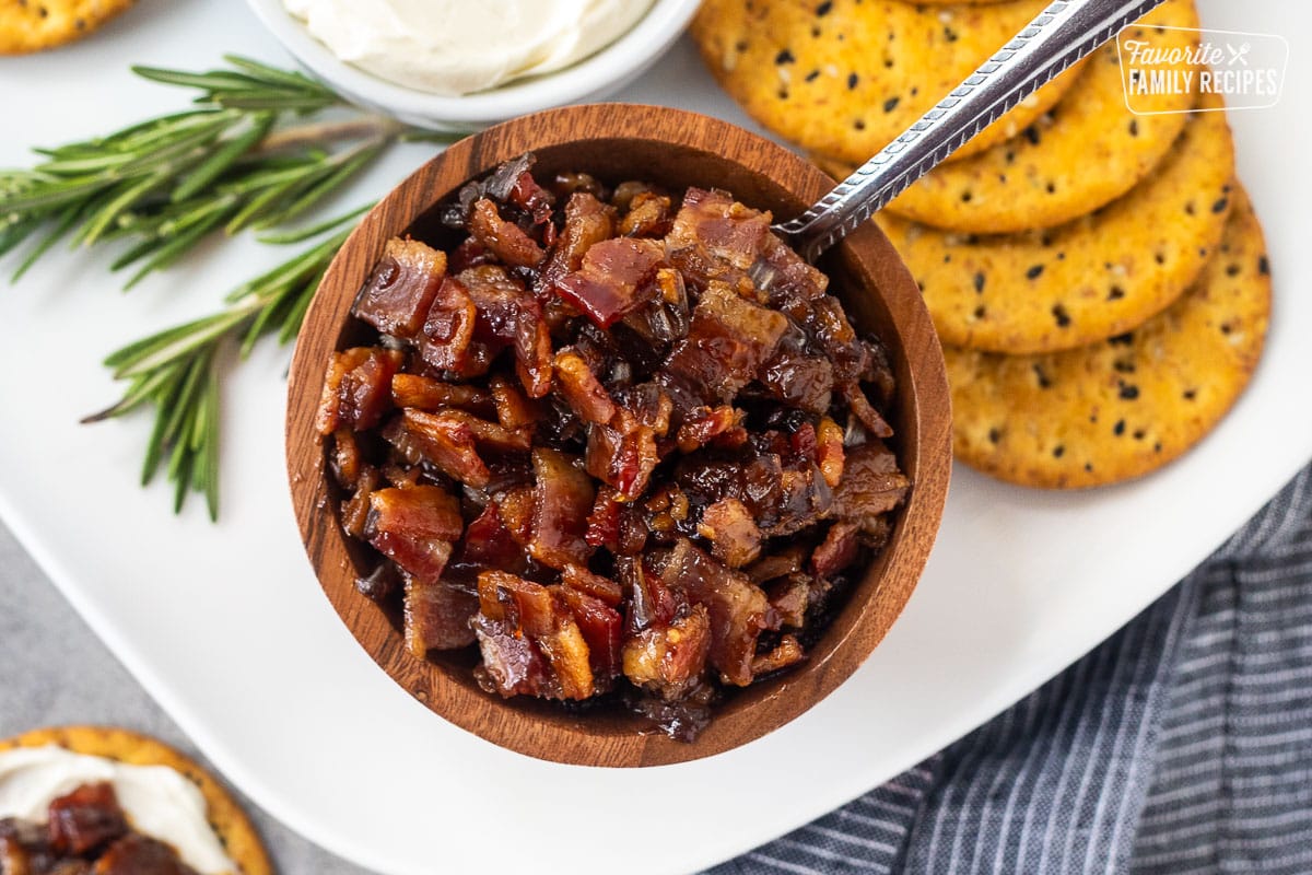 A wooden bowl full of bacon jam with a spoon in the bowl. The bowl is sitting on a white plate next to a stack of crackers and bowl of cream cheese.