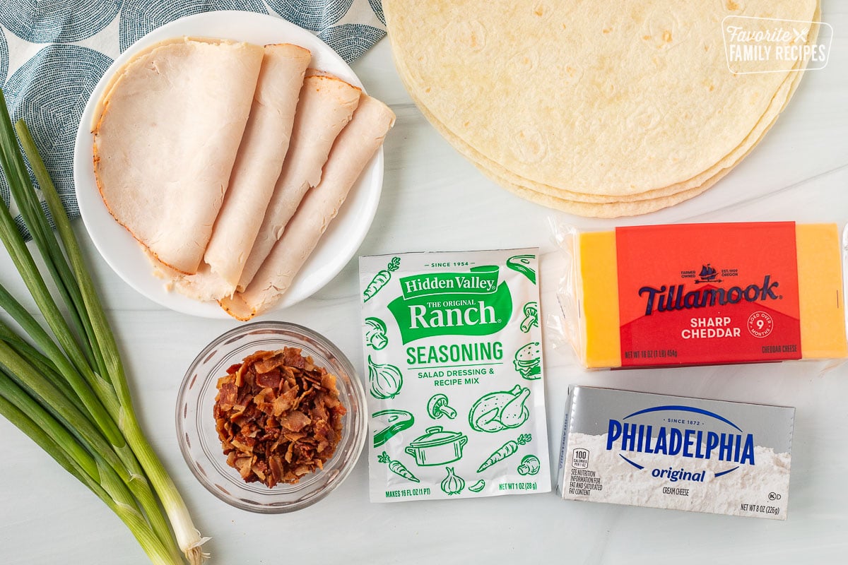 Ingredients to make turkey pinwheels including tortillas, sharp cheddar cheese, cream cheese, ranch seasoning, crumbled bacon, turkey slices, and green onions.