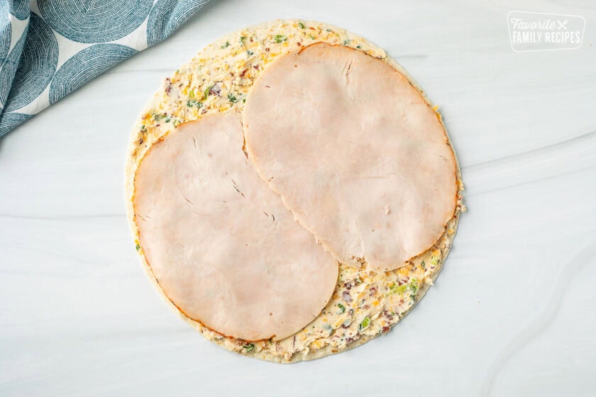 A tortilla laying flat on a surface with a spread of cream cheese and turkey slices over the top