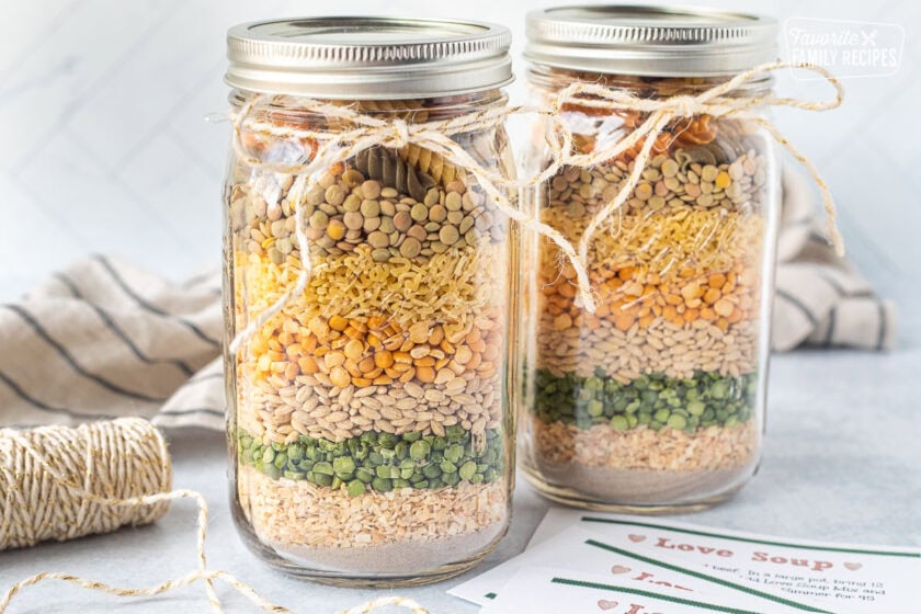 Two mason jars with layered ingredients to make soup including beef bouillon, dried onions, split green peas, barley, yellow peas, alphabet pasta, lentils and tri colored pasta with a string tied around and labels.