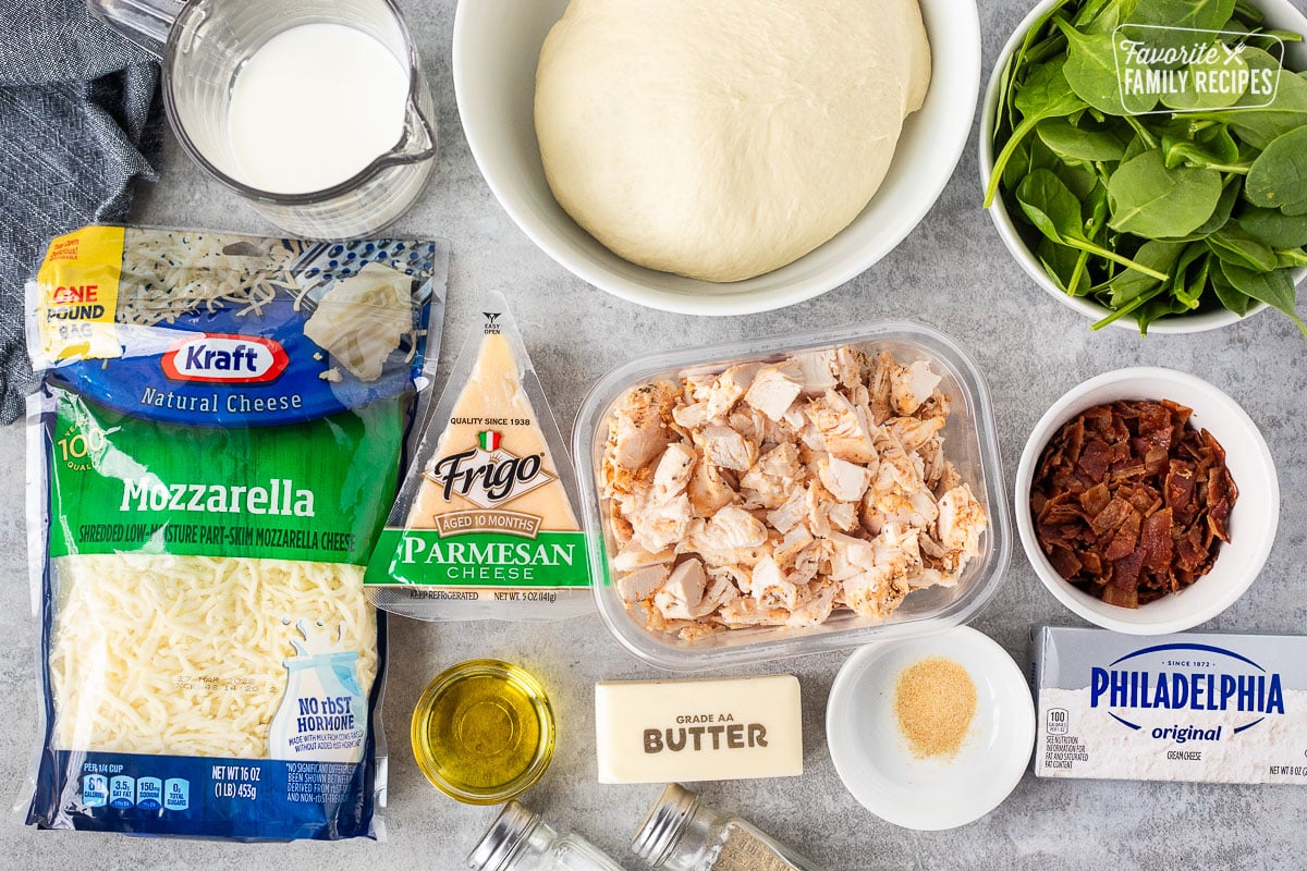 Ingredients to make Alfredo pizza including pizza dough, spinach, rotisserie chicken, bacon, cream cheese, garlic powder, butter, olive oil, salt, pepper, Parmesan cheese, mozzarella and milk.