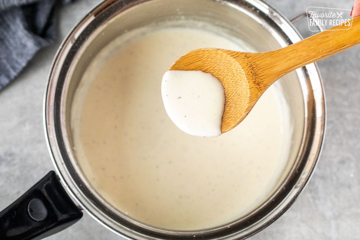 Wooden spoon holding up a scoop of Alfredo sauce over a saucepan.