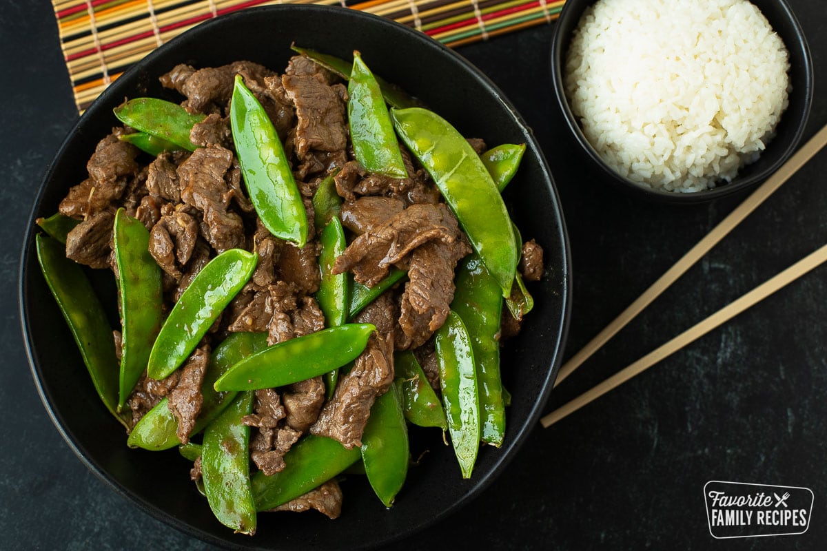 A serving bowl of beef and snow peas next to a small bowl of white rice and chopsticks