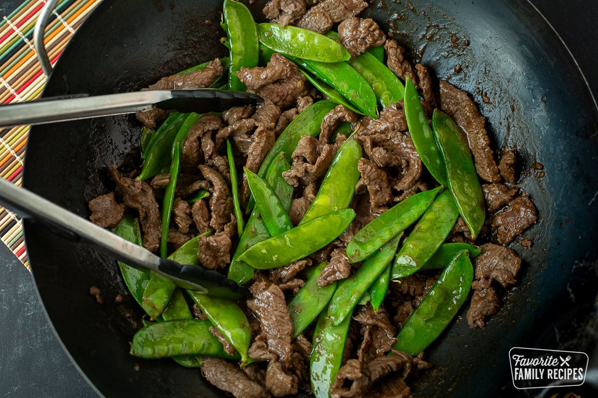 A close up of a wok of beef and snow peas with tongs tossing it in sauce