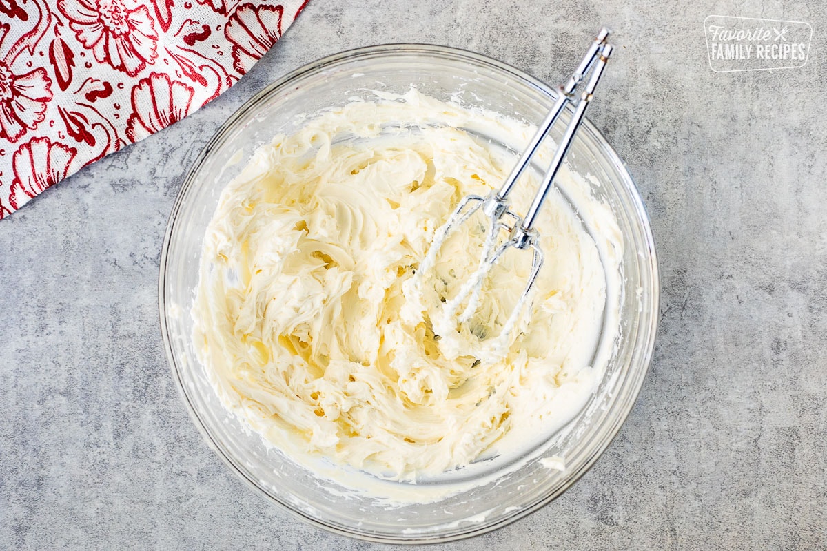 Glass mixing bowl with whipped cream cheese and electric beaters resting in the bowl.