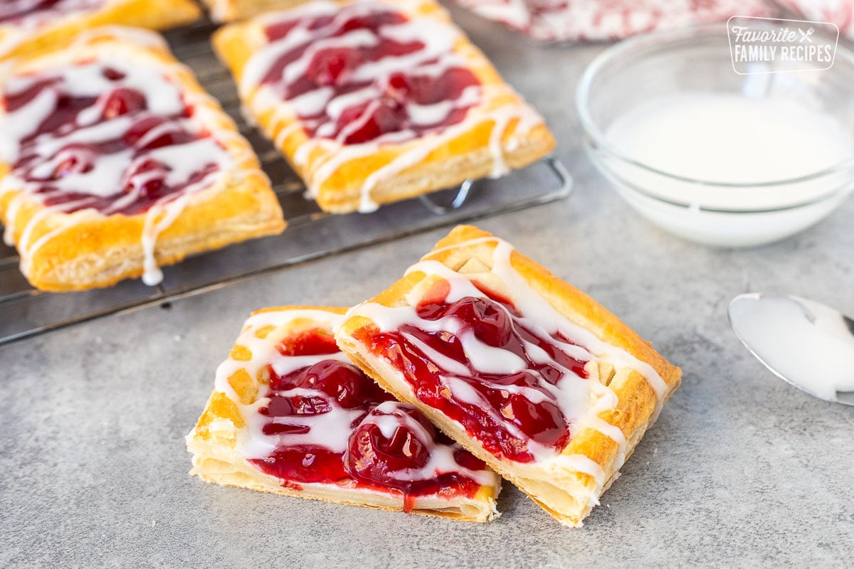 Cherry danish, cut and half with white icing. Cooling rack of cherry danishes in the background with a dish of white icing and spoon.