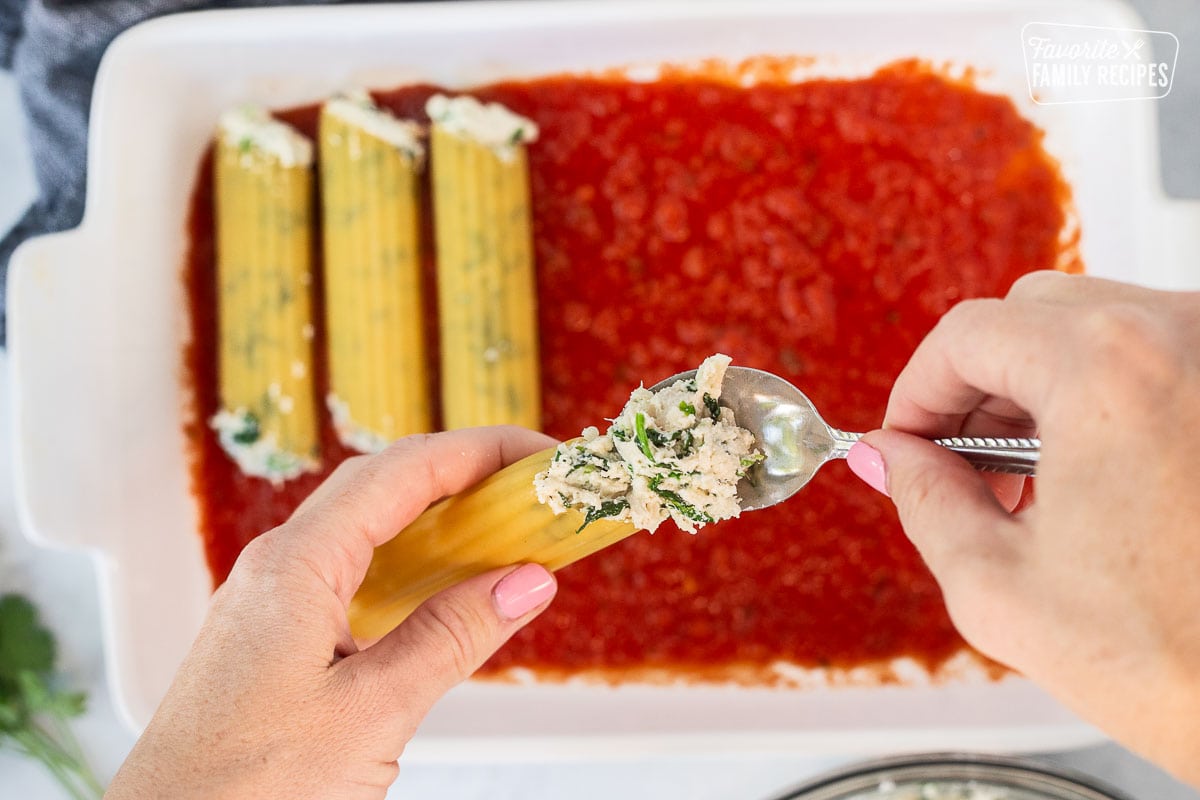 Filling dry manicotti shells with chicken and spinach filling using a spoon.