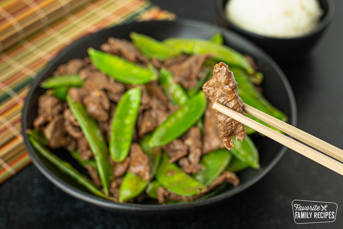 A close up of a slice of beef being help by chopsticks above a bowl of beef and snow peas