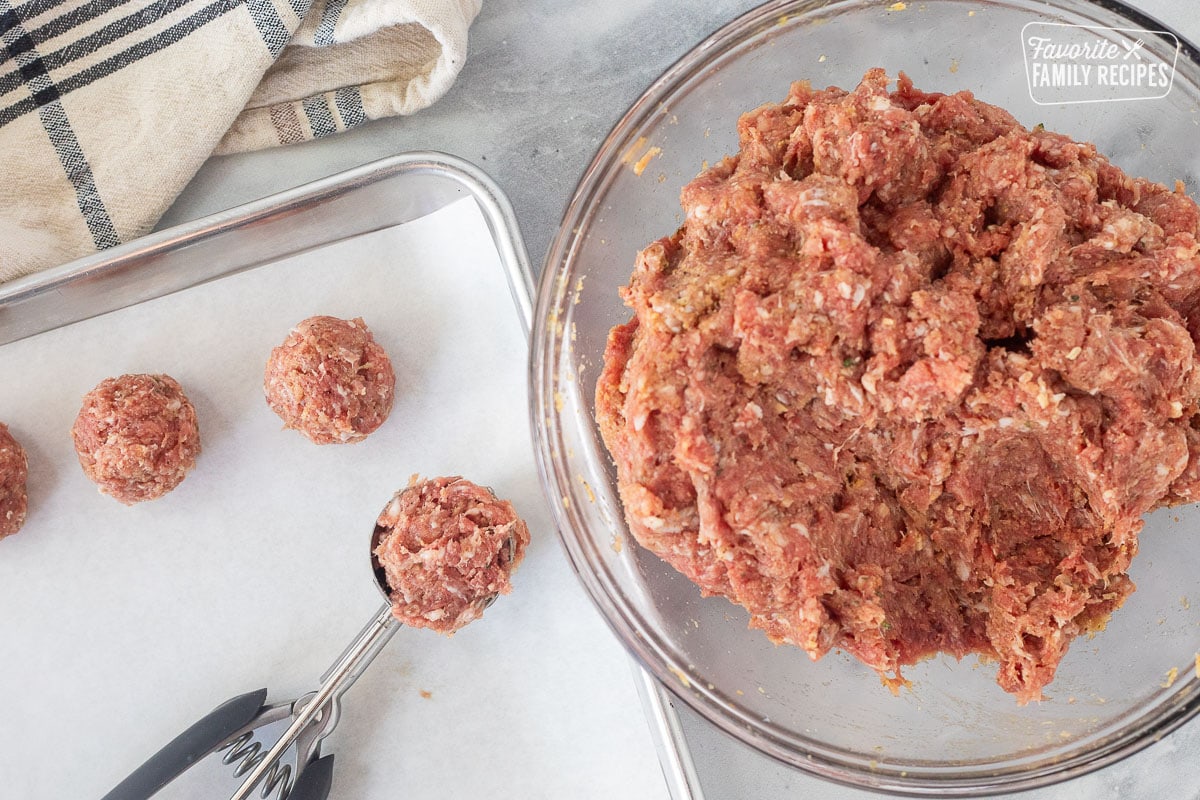 Scooped meatballs on a cookie sheet next to a bowl of meat mixture.