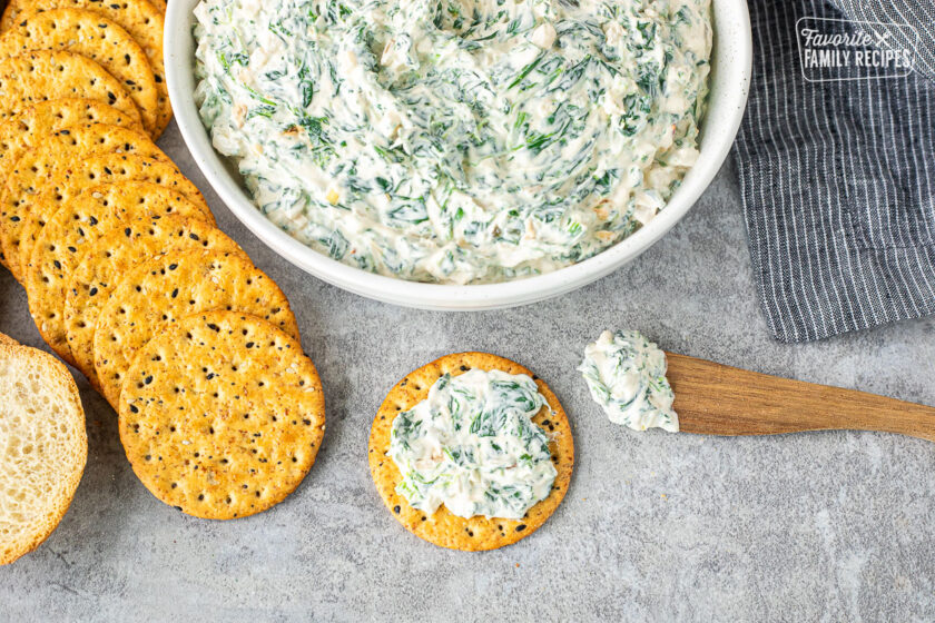 Spreading easy spinach dip on a cracker with a knife next to a large bowl of easy spinach dip.