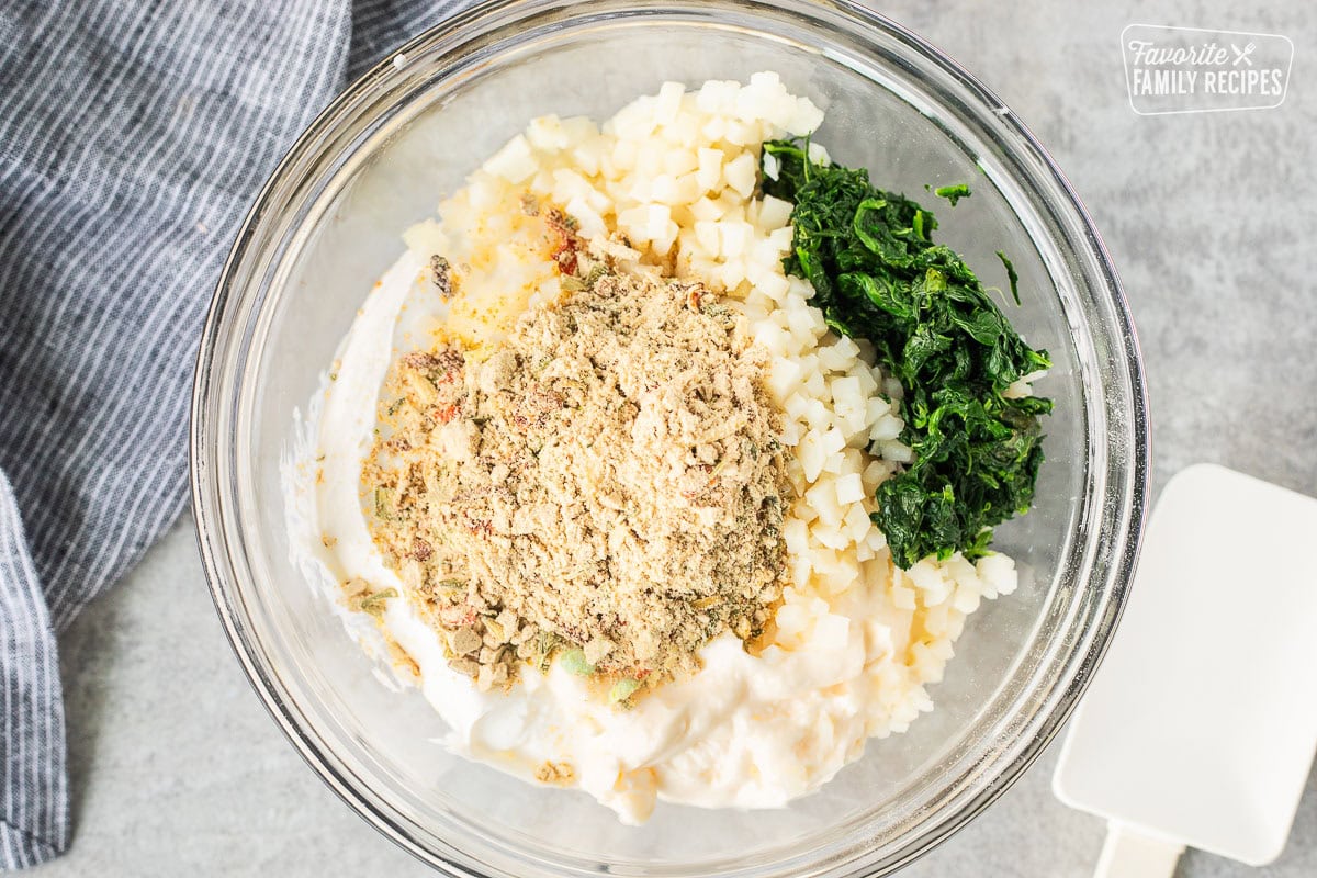 Glass mixing bowl with spinach, water chestnuts, Knorr vegetable mix, mayonnaise and sour cream. Spatula on the side.