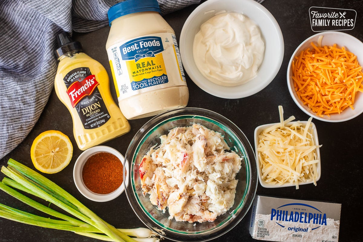 Ingredients to make Hot Crab Dip including crab, cheddar cheese, parmesan cheese, mayo, sour cream, cream cheese, dijon mustard, lemon, Old Bay seasoning and green onions.