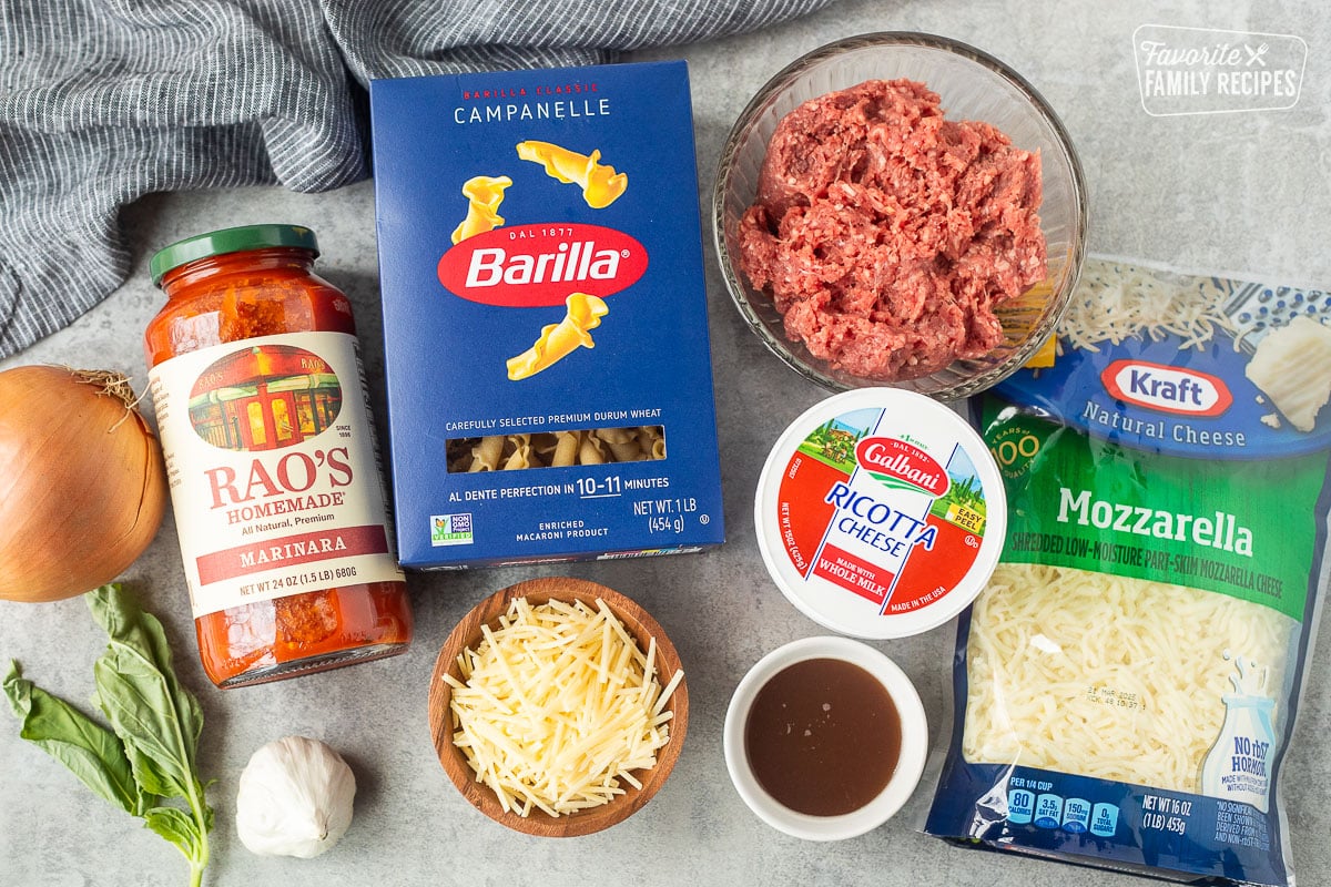 Ingredients to make lasagna casserole including ground beef, mozzarella cheese, ricotta cheese, beef broth, Parmesan cheese, Campanelle pasta, marinara sauce, garlic, onion and fresh basil.