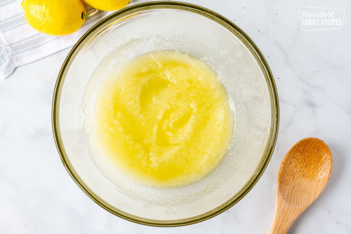 Mixing bowl of lemon juice and sugar to make Lemon Poppy Seed Muffin. Wooden spoon on the side.