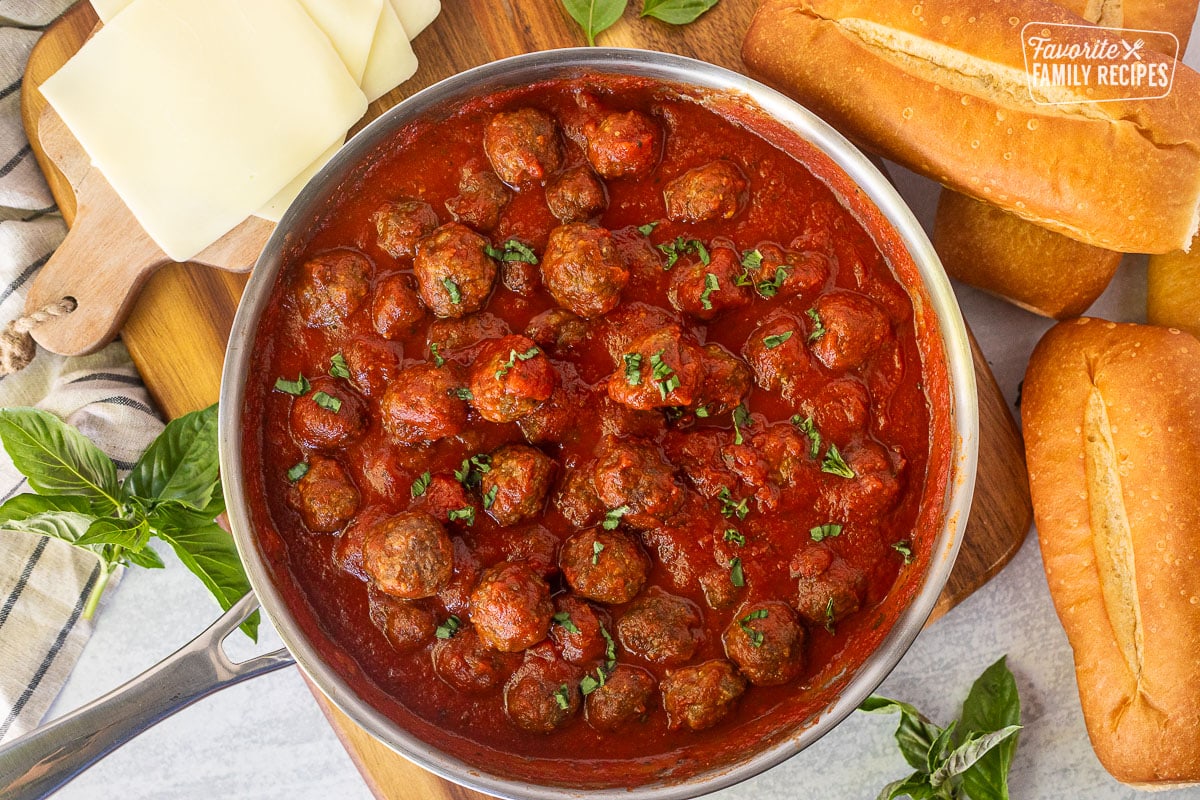 Meatballs in marinara sauce in skillet surrounded by rolls and cheese.