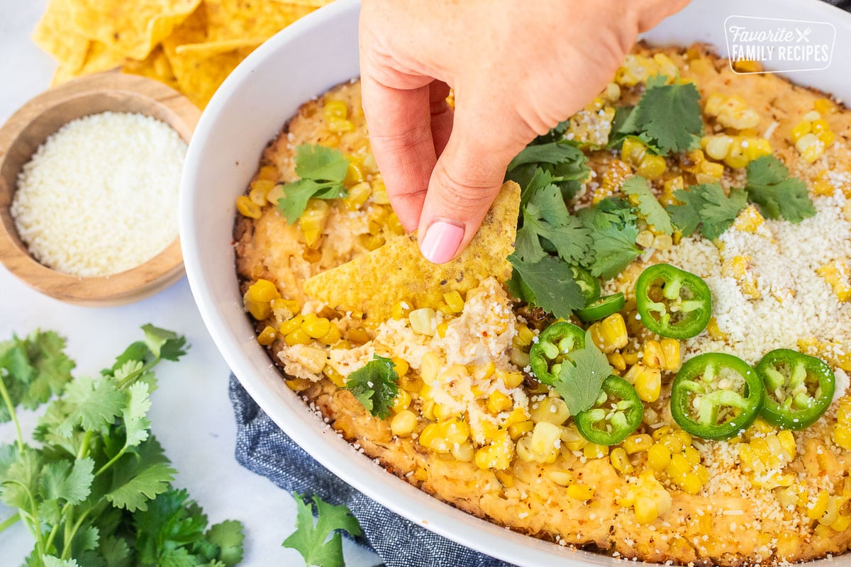 Hand dipping a tortilla chip into Mexican Street Corn Dip in a dish toped with jalapeño, cilantro and cojita cheese.