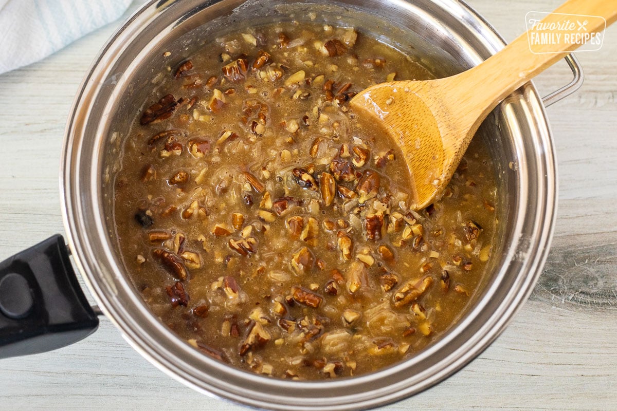 Saucepan with pecans and caramel added to brown sugar butter mixture with a wooden spoon resting in the pan.