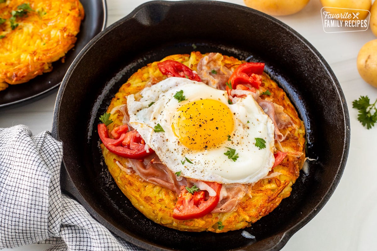 Rosti in a skillet topped with cheese, prosciutto, tomatoes, egg and parsley and rosti on a plate.