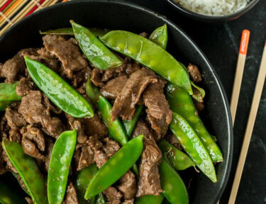 A vertical image of a serving bowl of beef and snow peas next to a small bowl of white rice and chopsticks