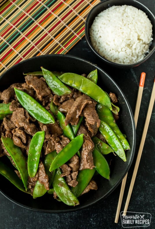 A vertical image of a serving bowl of beef and snow peas next to a small bowl of white rice and chopsticks