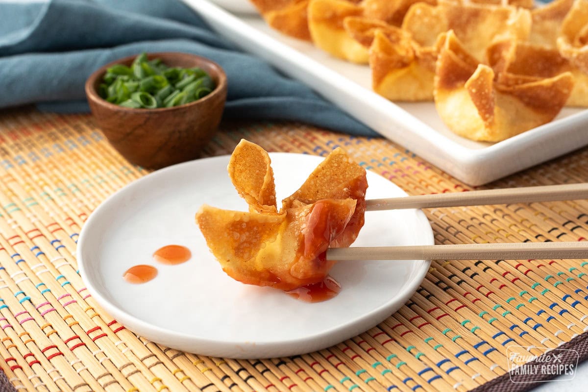 Close up of a fried, crispy cream cheese wonton being held with chopsticks and sweet and sour sauce dripping from it