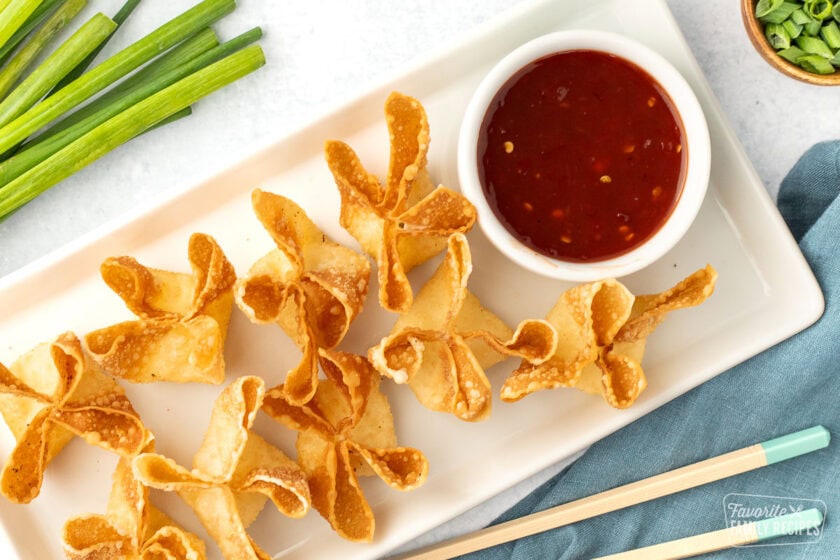 Top-down view of a plate of golden, fried cream cheese wontons next to a bowl of sweet and sour sauce