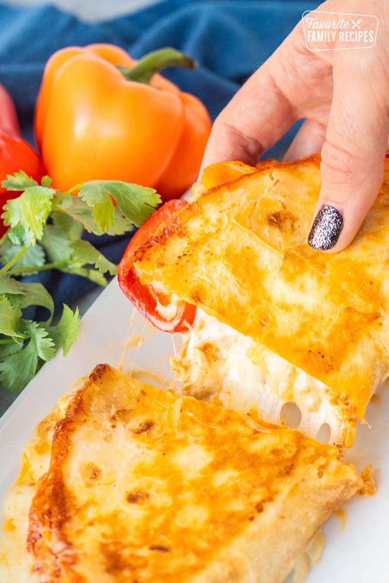 Hand lifting half of a chicken quesadilla on a plate with red and orange peppers.