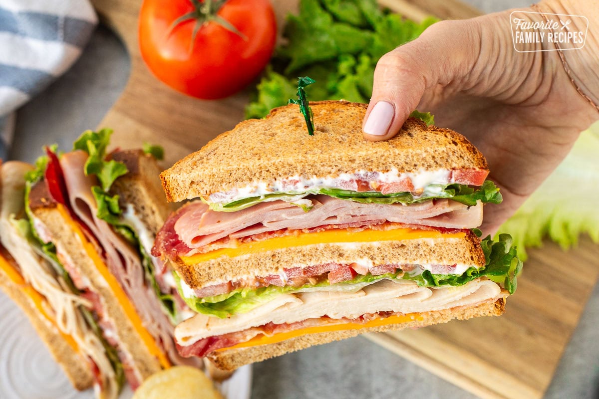 Hand holding a Club sandwich with mayo, cheddar cheese, bacon, turkey, lettuce, tomatoes and ham on toasted wheat bread. Potato chips on the side.