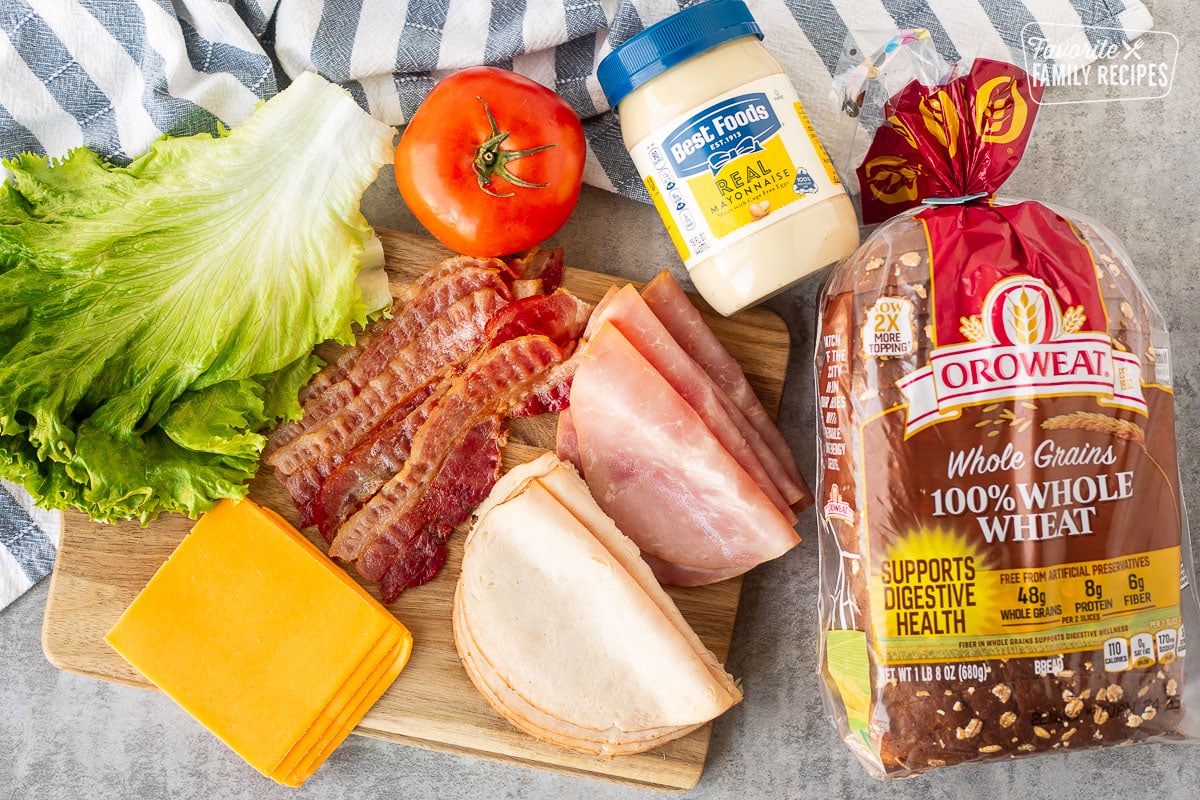 Ingredients to make club sandwich with wheat bread including wheat bread, turkey slices, ham slices, mayonnaise, tomato, bacon slices, cheddar cheese slices and lettuce.