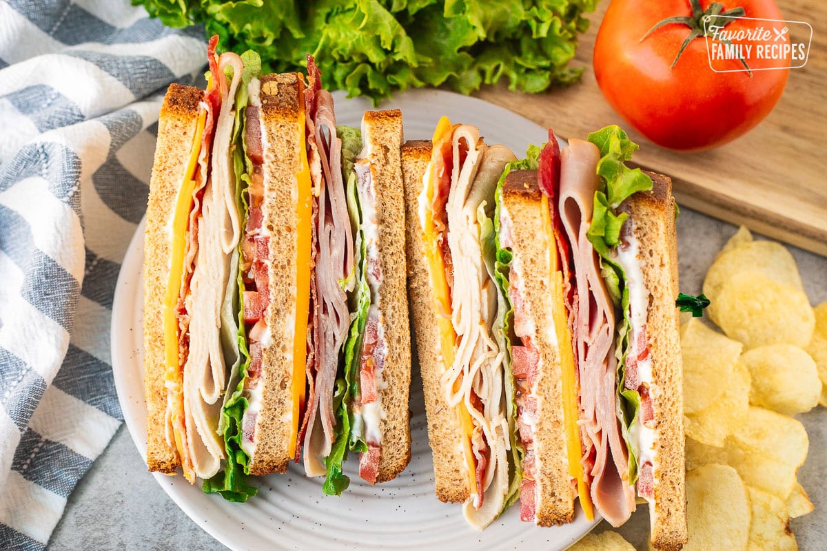 Club sandwich with mayo, cheddar cheese, bacon, turkey, lettuce, tomatoes and ham on toasted wheat bread. Potato chips on the side.