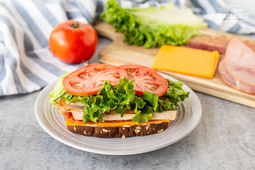 Play with slice of toasted bread with mayo, cheddar cheese, bacon, turkey, lettuce and tomato stacked on top. Cutting board with cheddar cheese, ham, bacon, lettuce and tomato tomato in the background.