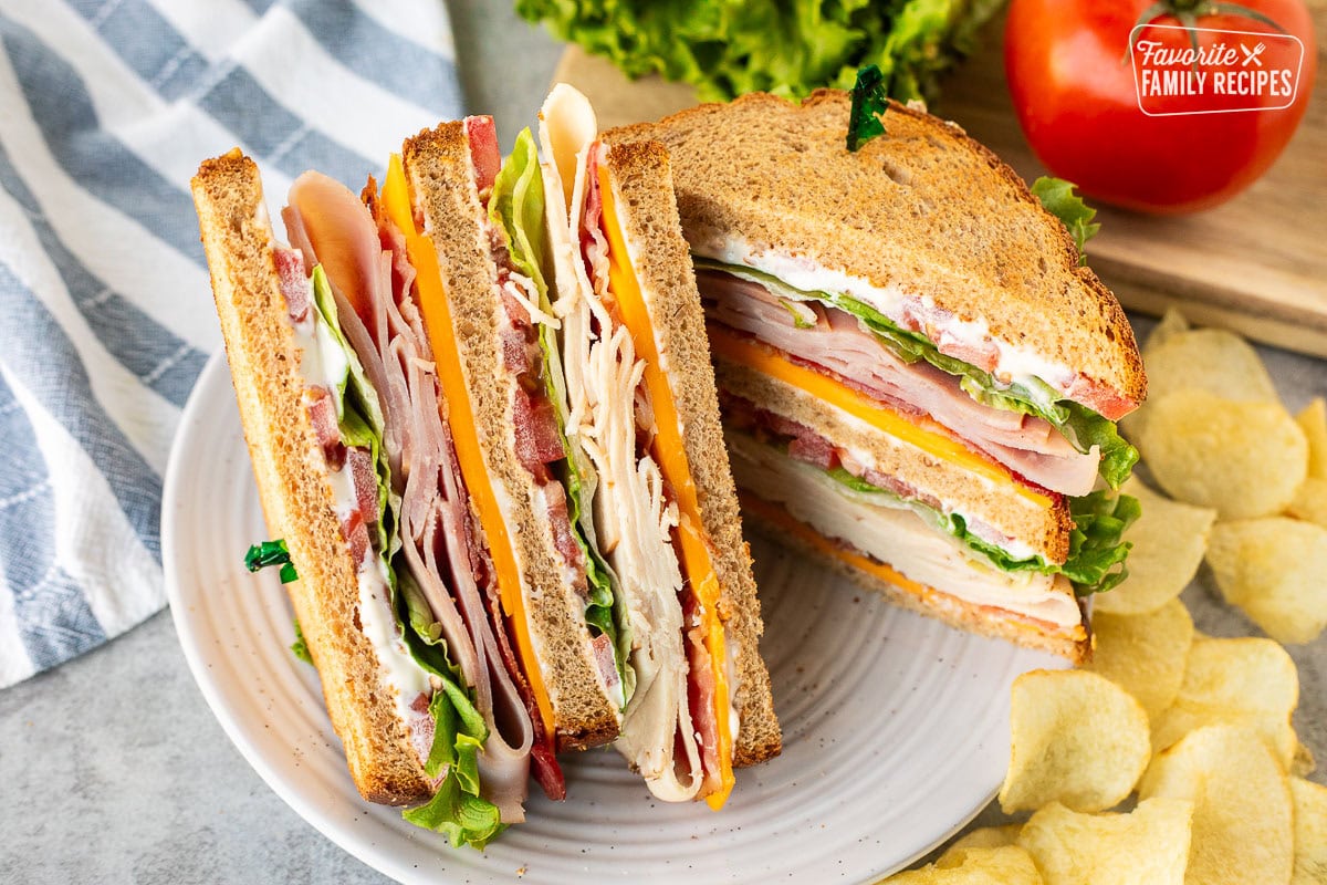 Club sandwich cut in half with mayo, cheddar cheese, bacon, turkey, lettuce, tomatoes and ham on toasted wheat bread. Potato chips on the side.