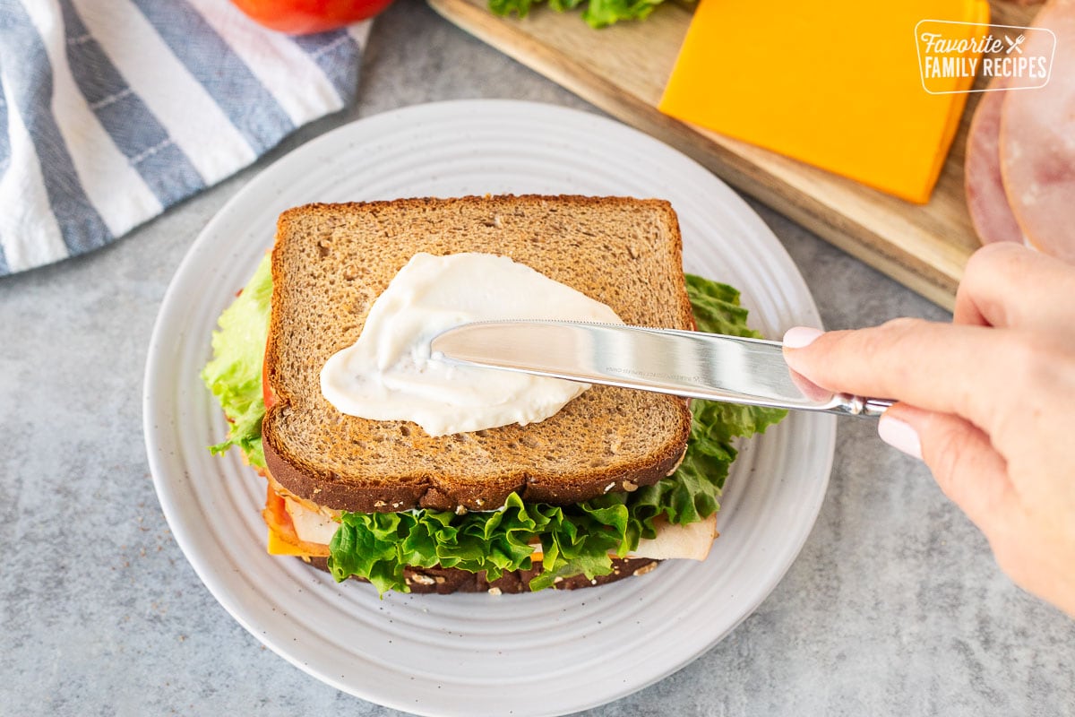 Spreading mayo with a knife on a slice of wheat toast on the top of a sandwich.