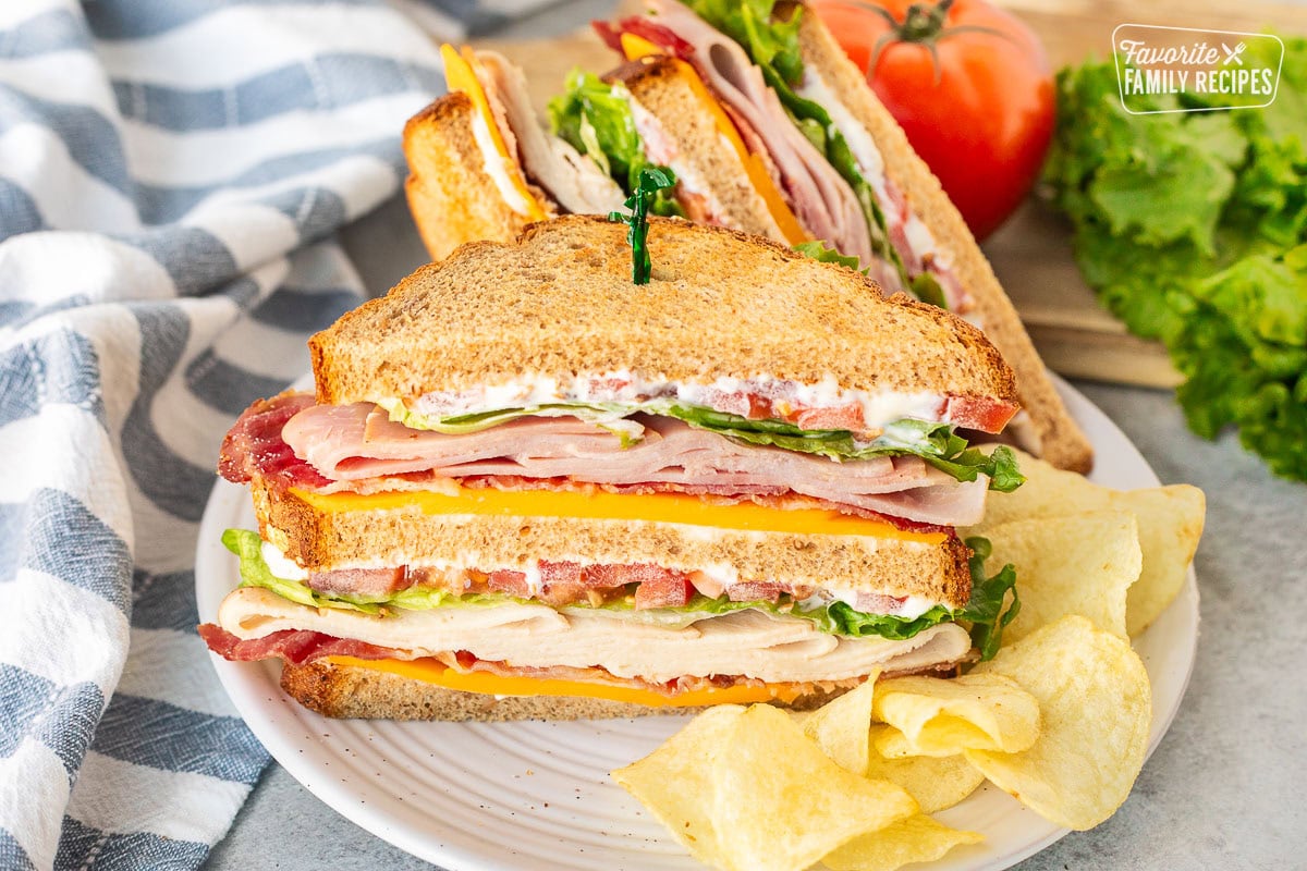 Club sandwich cut in half with mayo, cheddar cheese, bacon, turkey, lettuce, tomatoes and ham on toasted wheat bread. Potato chips on the side.