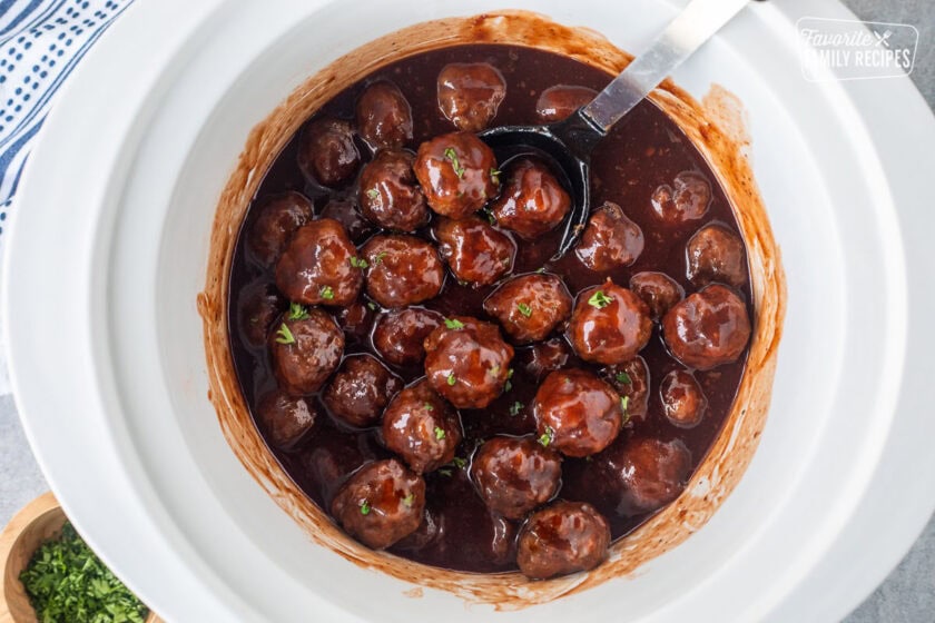 Crockpot of grape jelly meatballs with a spoon and garnished with fresh parsley.