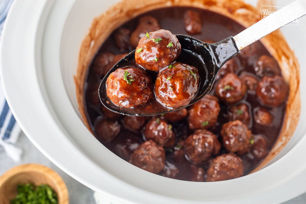 Spoon holding three grape jelly meatballs over a crockpot of grape jelly meatballs.