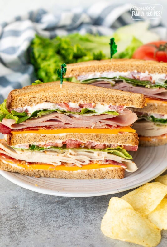 Club sandwich cut in half with mayo, cheddar cheese, bacon, turkey, lettuce, tomatoes and ham on toasted wheat bread. Potato chips on the side.