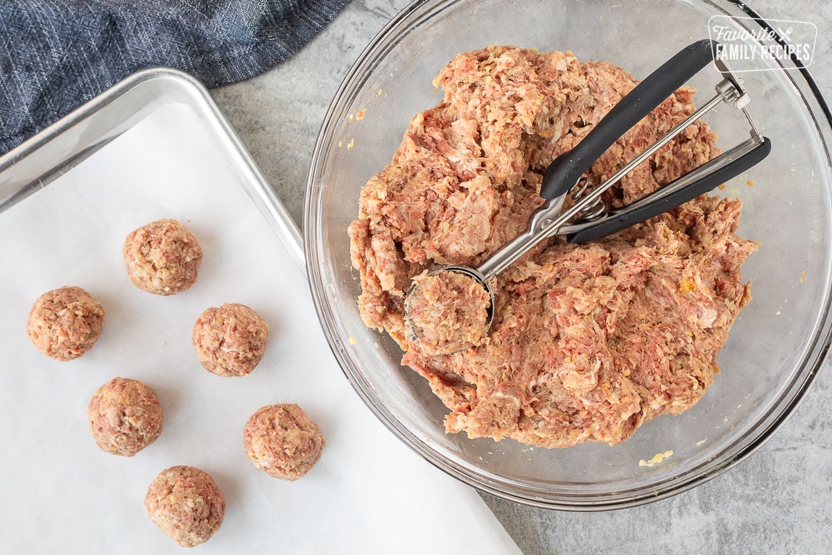 A cookie scoop scooping meat from a glass bowl with formed meatballs on a baking sheet ready to bake and freeze