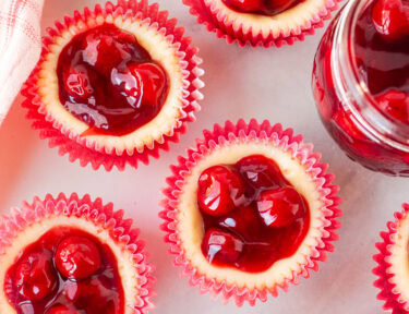 An overhead shot of six mini cherry cheesecakes next to a jar of cherries.