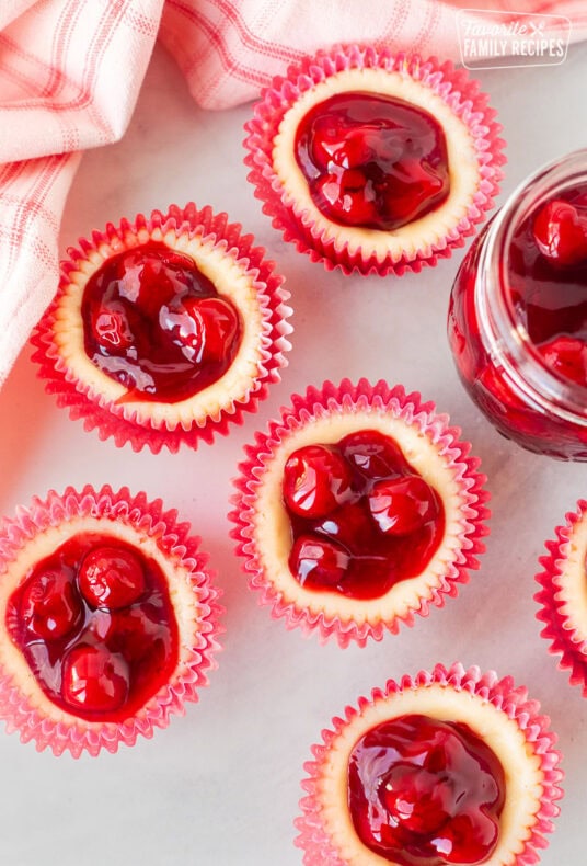 Individual mini cherry cheesecakes with cherry topping next to a jar of cherries.