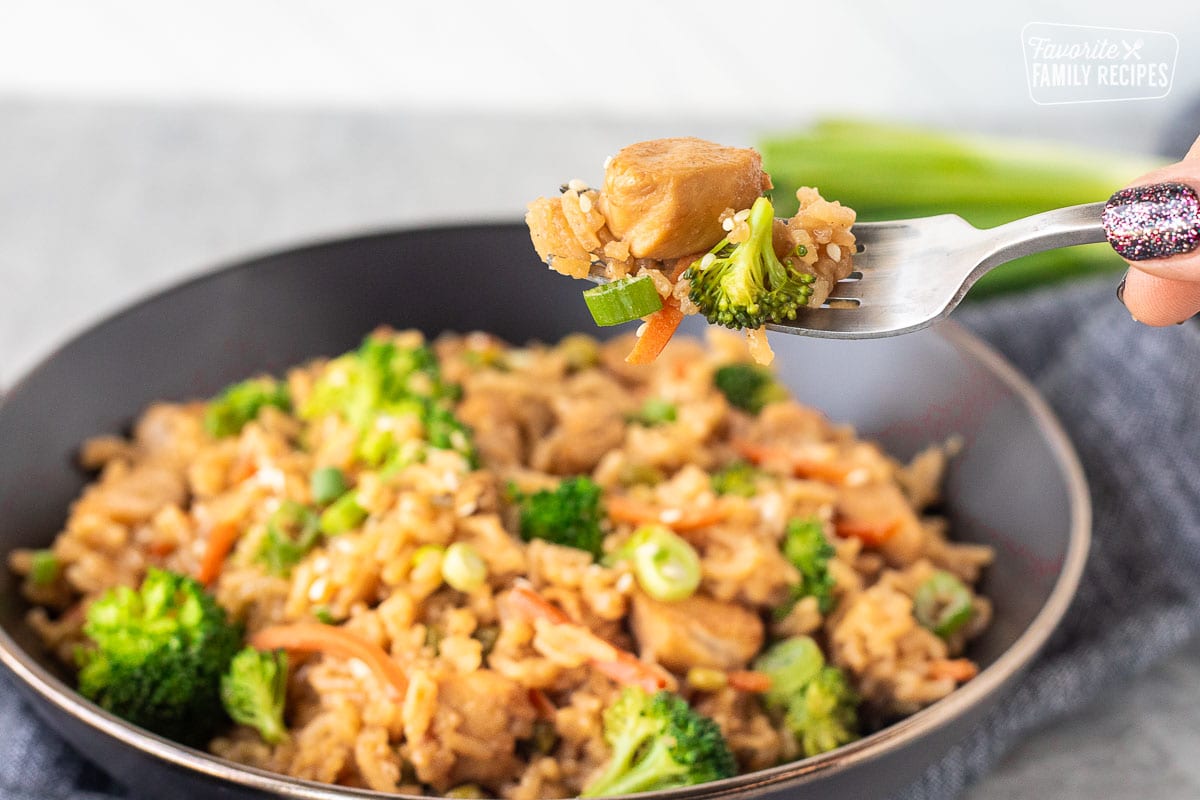  A fork lifting a bite of chicken teriyaki bowl with rice and vegetables from a dark bowl.