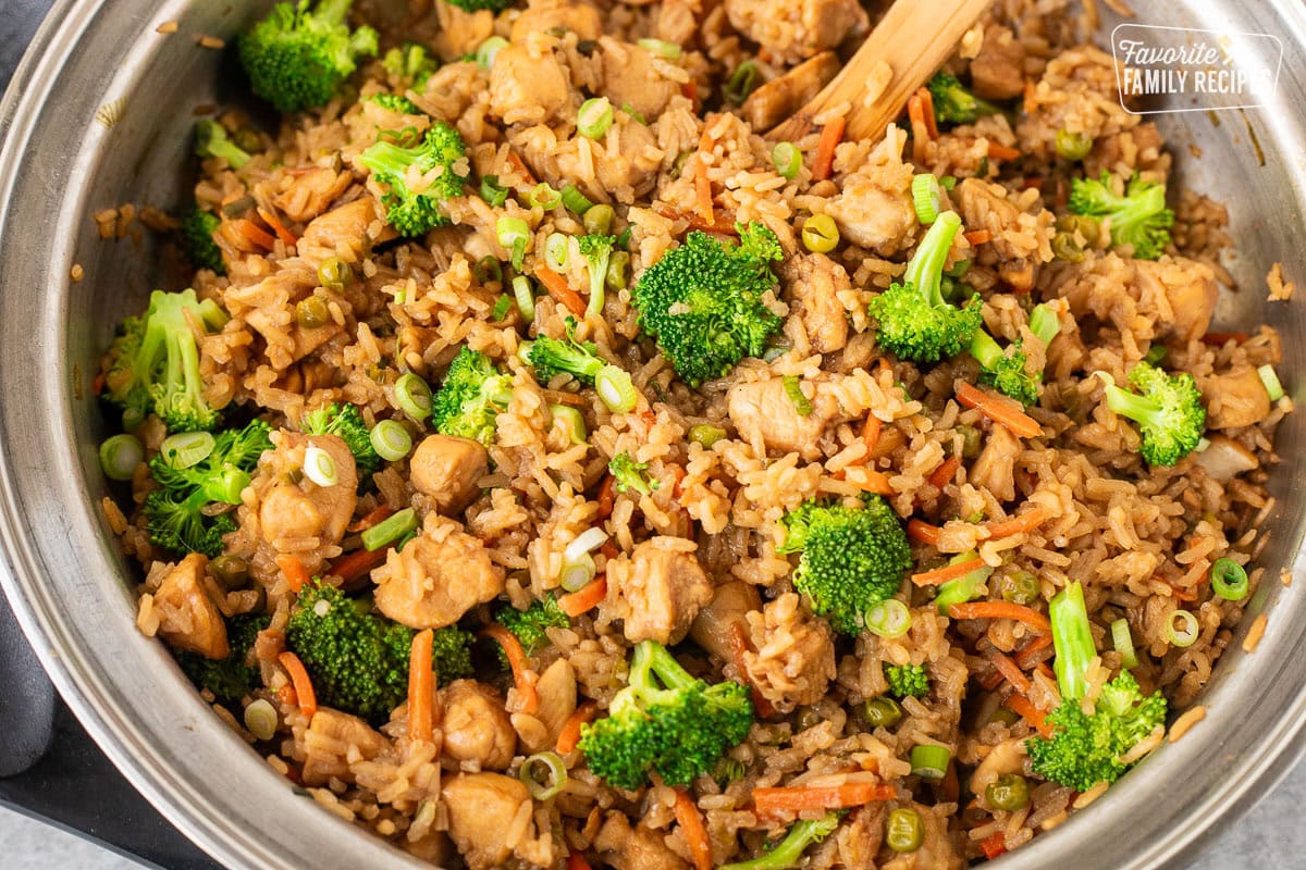 A stainless steel pan filled with teriyaki chicken, fried rice, broccoli florets, sliced carrots, and chopped green onions. A wooden spoon is visible, partially submerged in the rice. The pan is on a gray countertop.