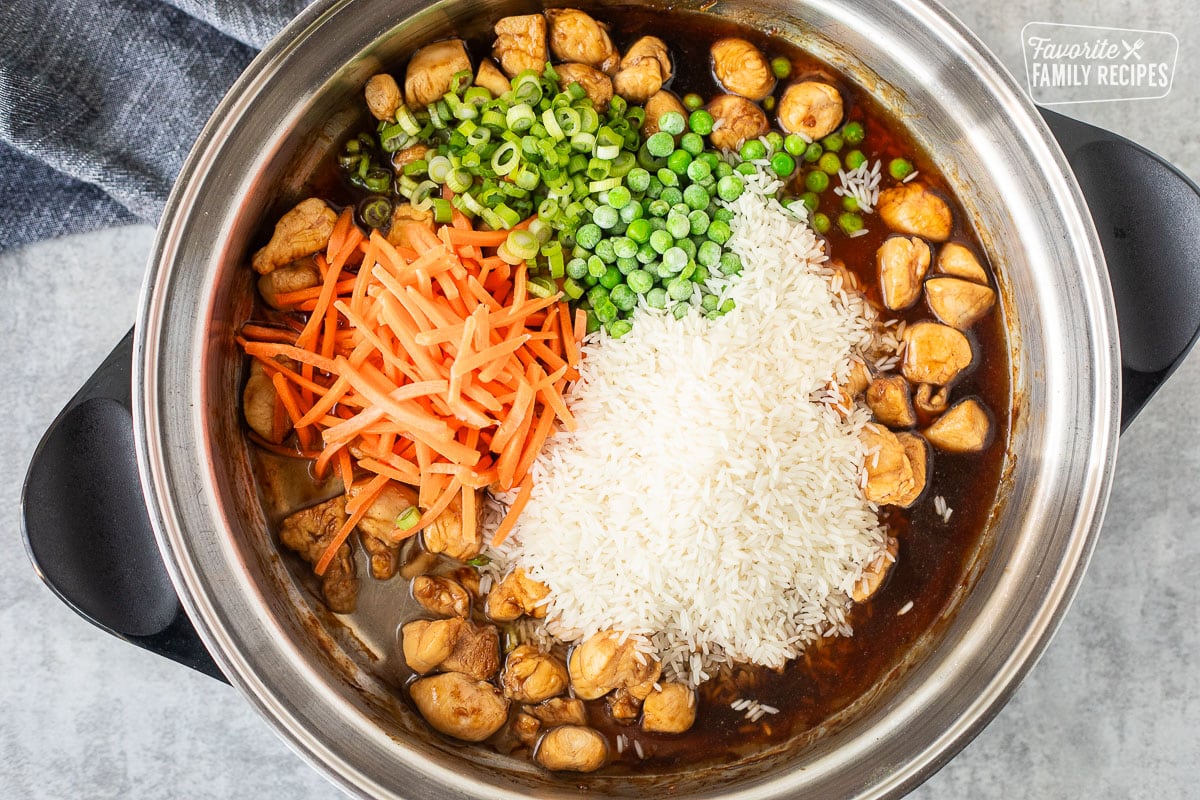 One pot teriyaki chicken bowl ingredients in a pan: chicken, rice, carrots, peas, and green onions.