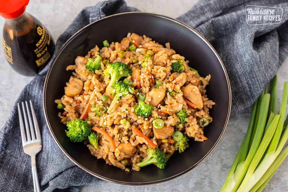 A dark bowl filled with teriyaki chicken and rice, featuring broccoli florets, sliced carrots, and chopped green onions. A bottle of soy sauce and a silver fork are placed to the left of the bowl, and a bundle of fresh green onions are on the right, all on a gray countertop with a gray cloth.