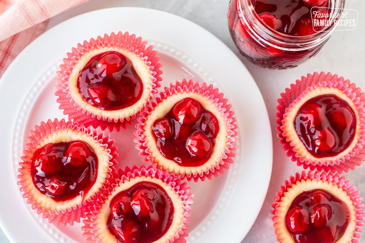 Mini cherry cheesecakes on a round plate next to a jar of cherries.