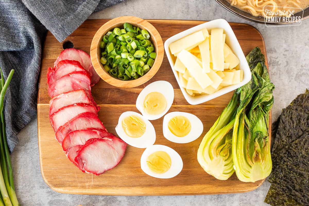 Ingredients to top Shoyu Ramen including barbecue pork loin, boiled eggs, bok choy, bamboo, green onion and seaweed sheets on a wooden cutting board.