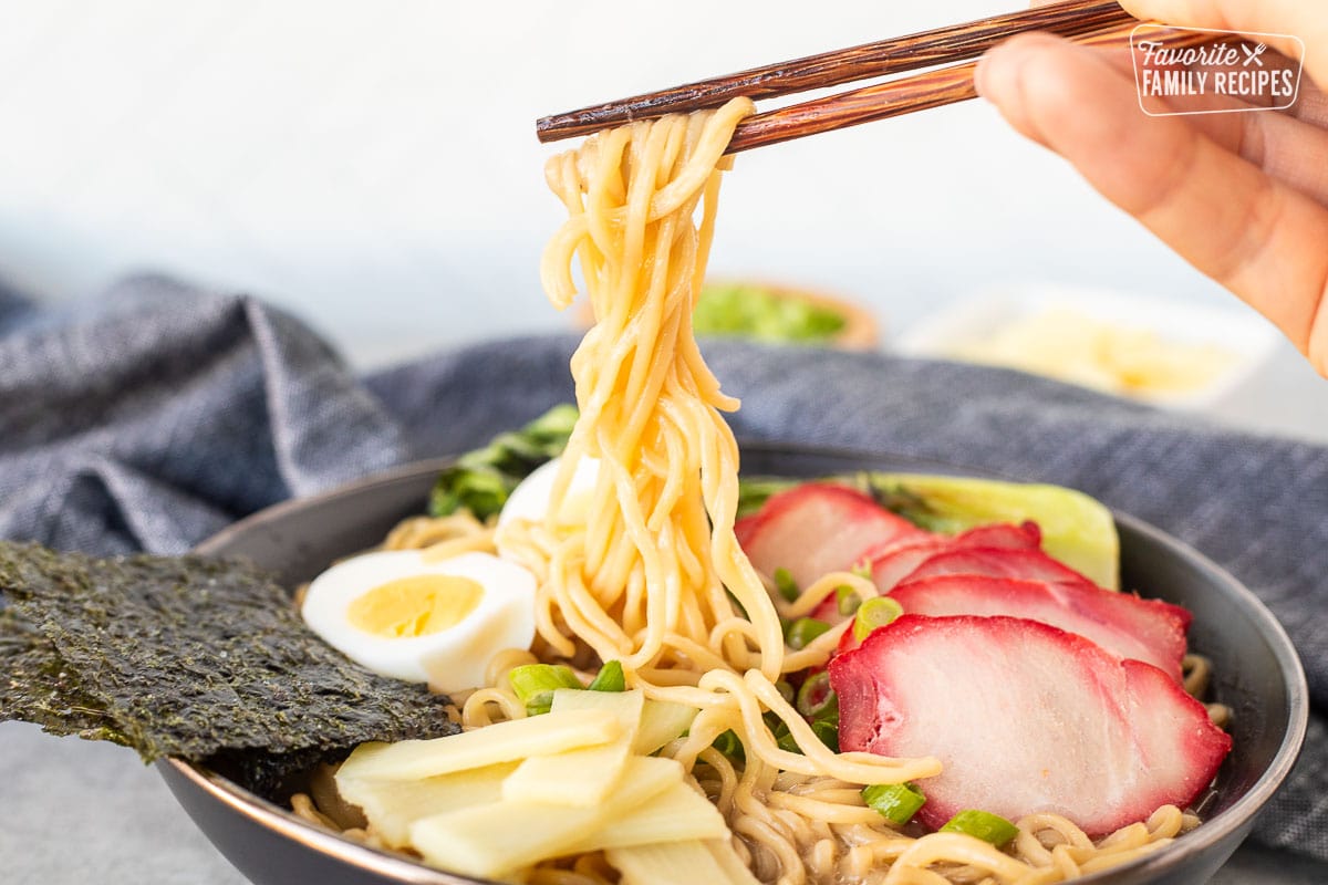 Wooden chopsticks holding up noodles from a bowl of Shoyu Ramen showing the delicious texture of the noodles.