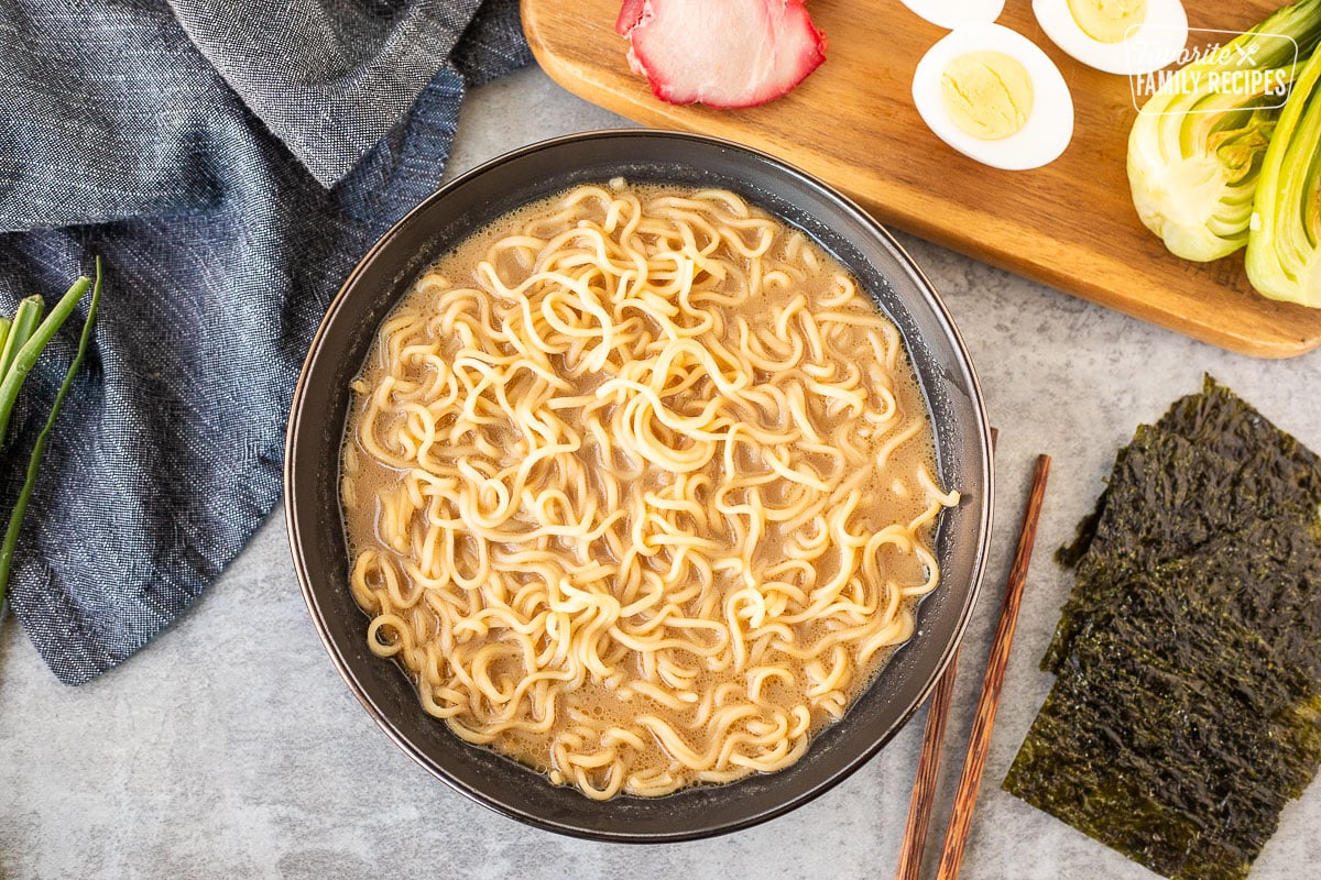 Bowl of Ramen broth and Ramen noodles next to seaweed sheets and a cutting board with barbecue pork loin, eggs and bok choy.