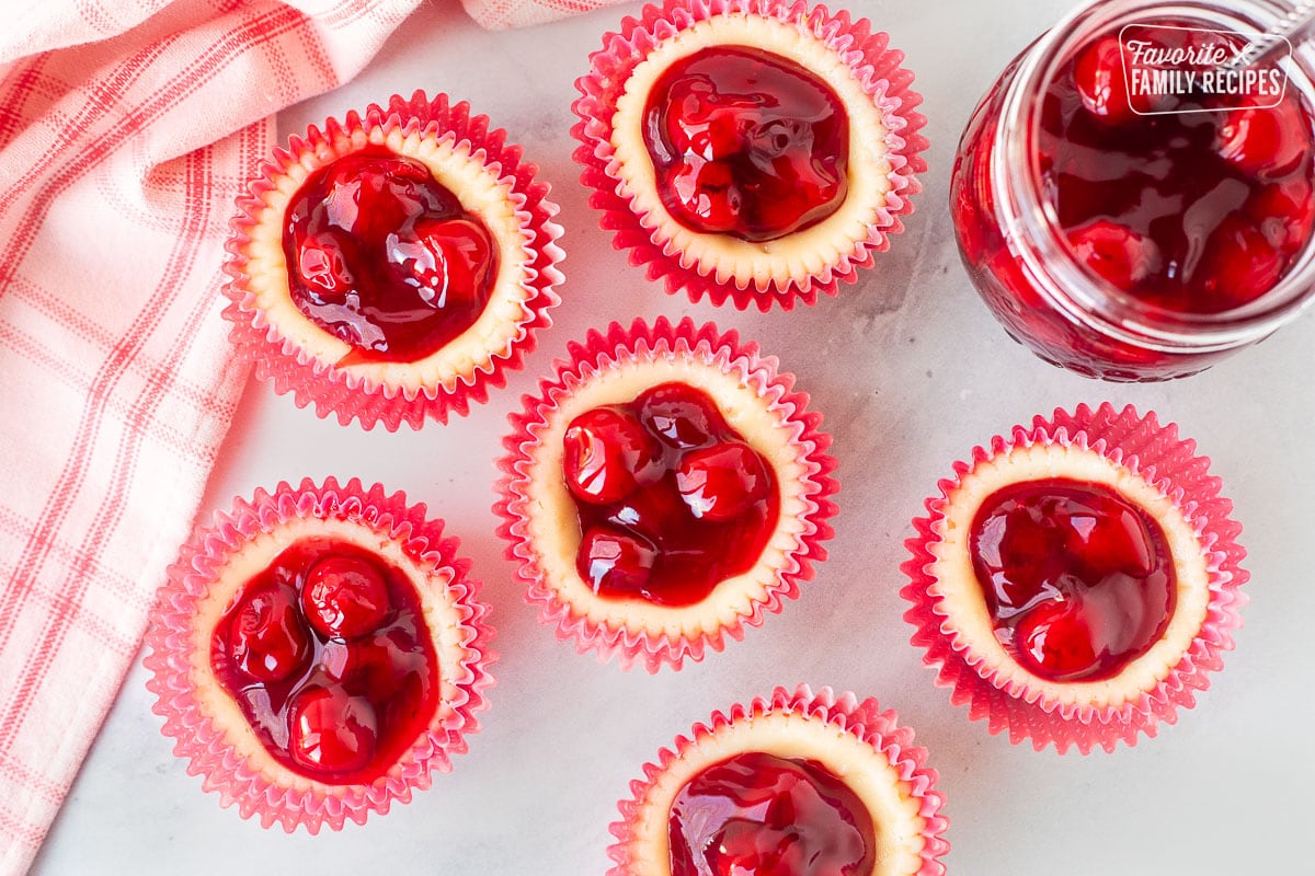 An overhead shot of six mini cherry cheesecakes next to a jar of cherries.